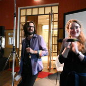 Andreas Scherffig und Hanna Hamel stehen vor einer Leinwand und halten Mikrofone in der Hand. Hanna Hamel klatscht.