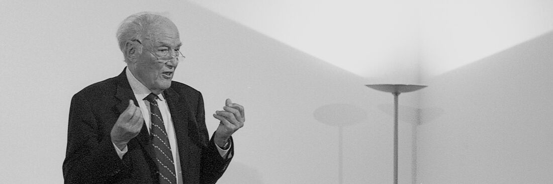 Eberhard Lämmert stands in front of a white wall, which is illuminated by a floor lamp on the right, and gesticulates as he speaks.