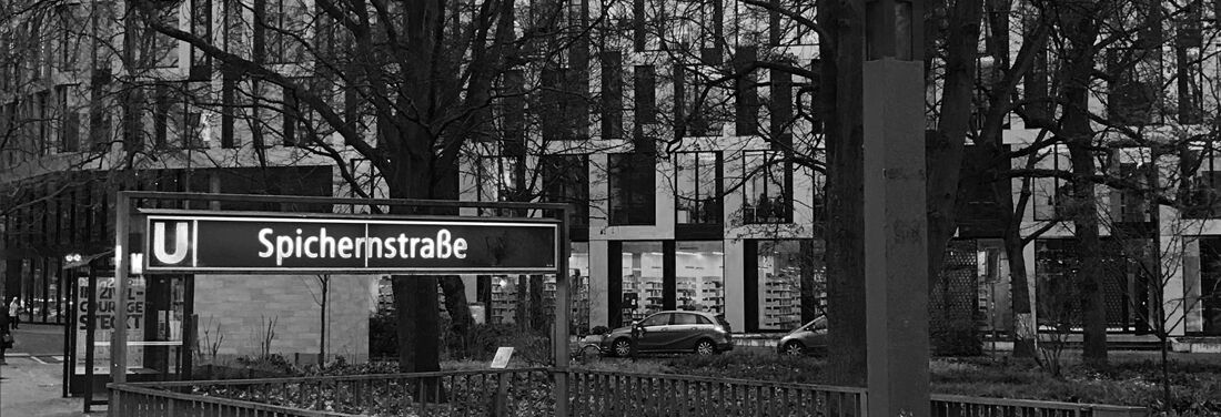 Schwarz-weiß-Bild vom U-Bahnhofeingang Spichernstraße am Rande der Gerhardt-Hauptmann-Anlage. Im Hintergrund ist der Neubau ACHTUNDEINS zu sehen, vor dem Autos parken.