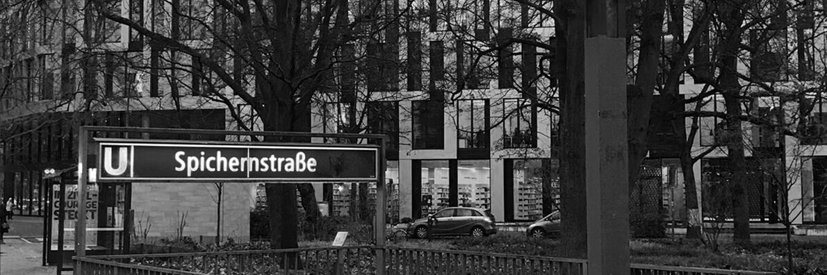 Black and white image of the Spichernstraße subway station entrance on the side of Gerhardt-Hauptmann-Anlage. The new ACHTUNDEINS building can be seen in the background, with cars parked in front of it.