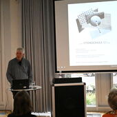 Ulrich Borgert stands at a bistro table with a laptop on it next to the lectern and speaks to the audience. Behind him is a presentation on the Itten School.