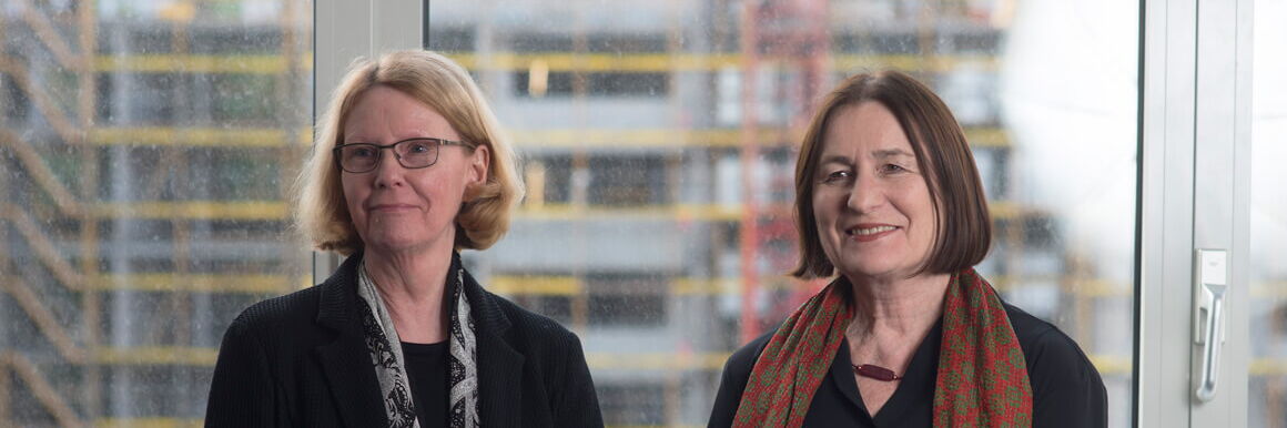 Franziska Thun-Hohenstein und Irina Scherbakowa stehen nebeneinander vor einem Fenster, hinter dem eine Baustelle zu sehen ist, und lächeln in die Kamera.
