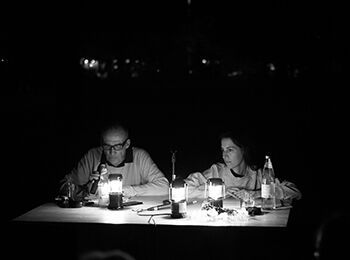 Tabea Hertzog und Cord Riechelmann sitzen an einem von kleinen Laternen beleuchteten Tisch im Freien. Um sie herum ist es dunkel.