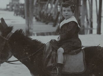 A black-and-white photo of a young boy wearing a cap and shorts, sitting on a donkey on the beach. A pier can be seen behind him.
