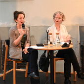 Anja Schenk and Martina Stemberger sit on a podium, Anja Schenk speaks into a microphone.