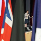 Frédérique Dreifuss-Netter steht am Pult und spricht in ein Mikrofon, im Vordergrund sind unscharf verschiedene Nationalflaggen zu erkennen.