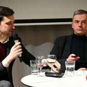 Lukas Laier und Christoph Hesse sitzen an einem Bistrotisch. Lukas Laier spricht in ein Mikrofon, Christoph Hesse hält eine Fernbedienung in der Hand.