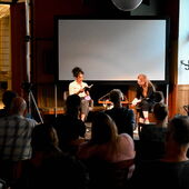 Elisa Aseva und Hanna Hamel sitzen auf einem Podium vor einer Leinwand, vor ihnen ist ein Publikum zu sehen. Elisa Aseva liest aus einem Buch vor.