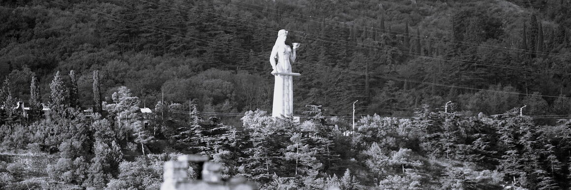 Schwarz-weiß-Foto einer großen weißen Frauenfigur mit einer Schale in der linken und einem Schwert in der rechten Hand. Um sie herum ist Wald.