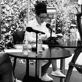 Jackie Thomae sitzt auf einer kleinen Bühne und liest aus einem Buch vor.