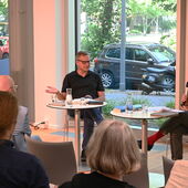 Georg M. Oswald und Patrick Hohlweck sitzen nebeneinander an weißen Bistrotischen und unterhalten sich. Vor ihnen sind die Hinterköpfe des Publikums zu sehen.