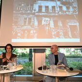 Katja Petrowskaja und Claude Haas sitzen nebeneinander an weißen Bistrotischen. Hinter ihnen ist ein historsiches Foto eines älteren Mannes zu sehen, der vor stark geschädigten Altbauten steht.