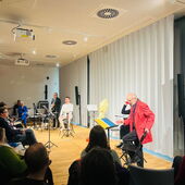 Several people are sitting on stools with music stands around them. In the background is a person with a saxophone, surrounded by many people sitting in the audience.