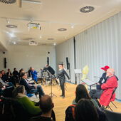 A person in black clothing with a black top hat declaims in front of an audience, surrounded by other actors and musicians.