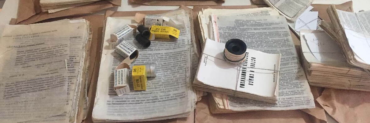 Foto eines Tisches, auf dem stapelweise eng bedruckte Papiere liegen. Auf den Papieren liegen kleine Röllchen mit Fotonegativen zusammen mit ihren Verpackungen und ein bedruckter, zusammengebundener Stapel Papier.