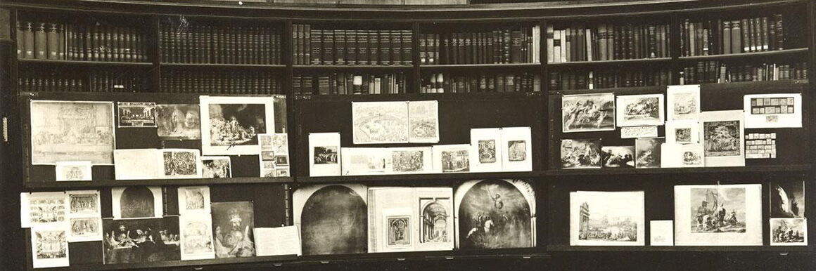 Schwarzweißaufnahme aus dem Lesesaal der Kulturwissenschaftlichen Bibliothek Warburg (KBW). Vor den Bücherregalen sind drei große schwarze Bildtafeln aufgestellt, an die jeweils mehrere verschieden große Abbildungen gepinnt sind.