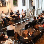 The speakers sit on a podium in front of an audience in a courtyard. Hanna Hamel speaks into a microphone.