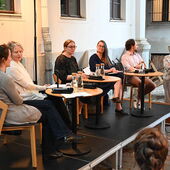 The speakers sit on a podium in front of an audience in a courtyard. Hanna Hamel speaks into a microphone.
