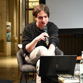The author Elias Hirschl sits at a laptop and speaks into a microphone.