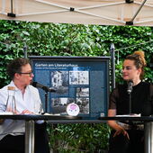 Eva Geulen und Teresa Präauer sitzen auf einer Bühne und unterhalten sich. Hinter ihnen ist eine Infotafel zum Garten am Literaturhaus zu sehen.