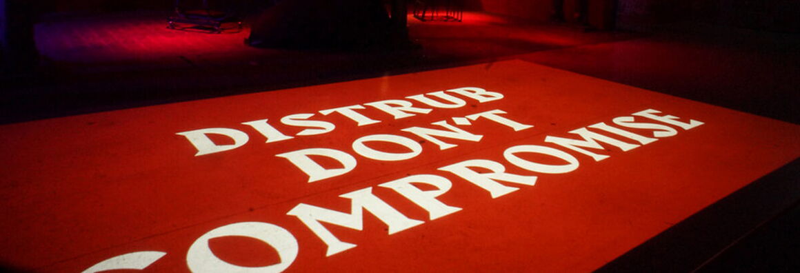 In einem dunklen Raum ist auf rotem Grund in weißen Lettern der Schriftzug "Distrub don't compromise" zu lesen.
