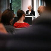 Sigrid Weigel steht am Rednerpult. Im Vordergrund sind verschwommene Publikumsreihen.