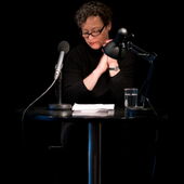 Eva Geulen sits at a table with microphone and lamp in front of a black background and looks down at a sheet of paper