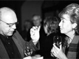 Black and white photograph of xxx and Angelika Neuwirth. In the foreground the two are talking with a champagne glass in their hands. In the background an event is going on..