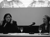 Black and white photograph of Rineke Dijkstra and xxx. The two are sit at a speaker's table. Rineke Dijkstra speaks into a microphone, xxx watches her.
