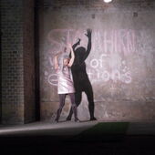 Eine Frau mit langem weißem Haar und glitzerndem Oberteil tanzt vor einer beschriebenen Betonwand, dabei wirft sie einen dunklen Schatten an die Wand