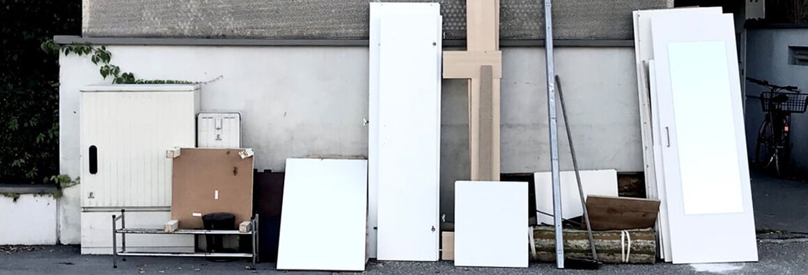 Verschiedene Möbelteile stehen an eine Wand gelehnt auf einem Bürgersteig an einer Straße mit Gulli.