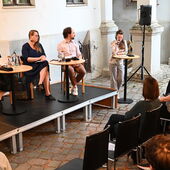 The speakers sit on a podium in front of an audience in a courtyard. Hanna Hamel speaks into a microphone.