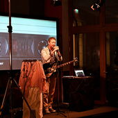 Musician Manu Louis stands on a stage and sings into a microphone. He carries a guitar and behind him on a large screen you can see the picture of a cassette and the text “Studio Master Cassette” written in the bottom left corner of the screen.