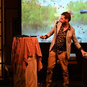 The musician Manu Louis is standing on a stage. In front of him is a microphone and he turns to the left to a high table with a white and red checkered tablecloth and electronic devices. With his left hand he touches a mixing desk.