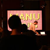 In the foreground are outlines of backs of heads. In the background, the musician Manu Louis is standing behind a mixing desk, and behind him the words “Manu Louis” are projected onto a screen in yellow letters on a red and white checkered background.