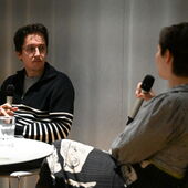 Jona Spreter and Ivana Sokola sit facing each other at a bistro table. Both are holding microphones, Sokola is speaking with her back to the viewer.