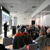 Detlev Schöttker stands at the lectern and speaks to the audience sitting in a large semicircle in front of him. There are around 60 people.