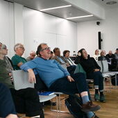 Several people sitting on gray chairs look forward with interest, smiling and even laughing.