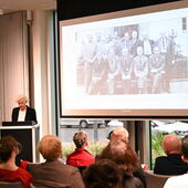 Sigrid Weigel steht am Rednerpult. Auf der Leinwand neben ihr ist ein schwarz-weißes Gruppenfoto von Männern in Anzügen zu sehen.