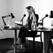 Schwarz-Weiß-Fotografie von Kat Kaufmann. Die Schriftstellerin Kat Kaufmann sitzt während einer Lesung auf einem Stuhl und gestikuliert mit der linken Hand.
