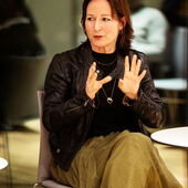 Anna A. Ahlers sitzt auf einem Stuhl vor einem bodentiefen Fenster und spricht, nach rechts gewendet und gestikulierend.