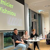 Zmicier Vishniou, Tatsiana Kupreichyk, and Nina Weller sit in front of the screen and look to the left, with the audience visible to their right.