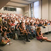 A full lecture hall, to the left of the rows of chairs people are sitting on the stairs and on the floor.