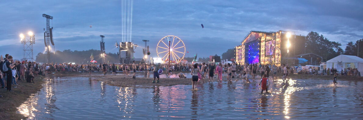 Scene from the Przystanek Woodstock festival. A large puddle can be seen in the evening light, with people dancing and playing. A brightly lit stage can be seen at the back right and a Ferris wheel in the middle.