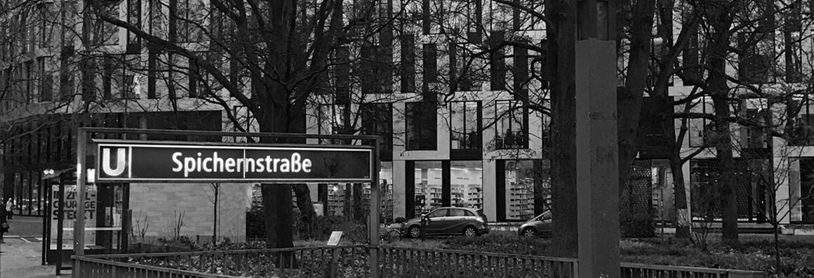 Schwarz-weiß-Bild vom U-Bahnhofeingang Spichernstraße am Rande der Gerhardt-Hauptmann-Anlage. Im Hintergrund ist der Neubau ACHTUNDEINS zu sehen, vor dem Autos parken.