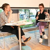 Kenah Cusanit und Hanna Hamel sitzen an weißen Bistrotischen und unterhalten sich.