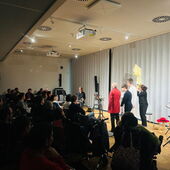 In front of full rows of spectators, four actors stand with their backs to the person watching and look at a person holding a saxophone.