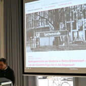 Eva Geulen stands at the lectern and speaks, with the title of the event and a black and white image of Spichernstraße subway station projected onto a screen behind her.