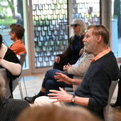 A man in a blue sweater and wristwatch sits in the audience and speaks.