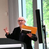 Detlev Schöttker stands behind the lectern and gesticulates as he speaks. He holds a pen in his right hand, a book and a yellow cardboard folder in his right.